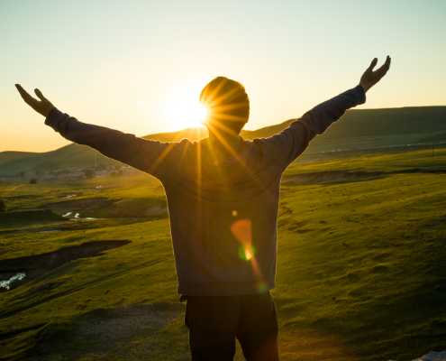 man standing on hill with arms out wide looking at sunset